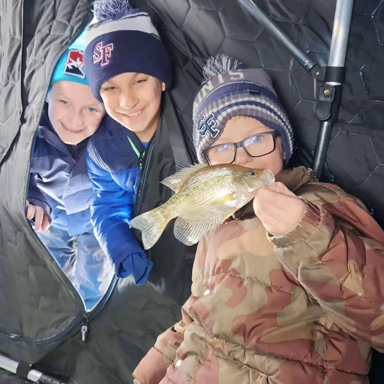 Three kids in an icehouse, one holding up a crappie