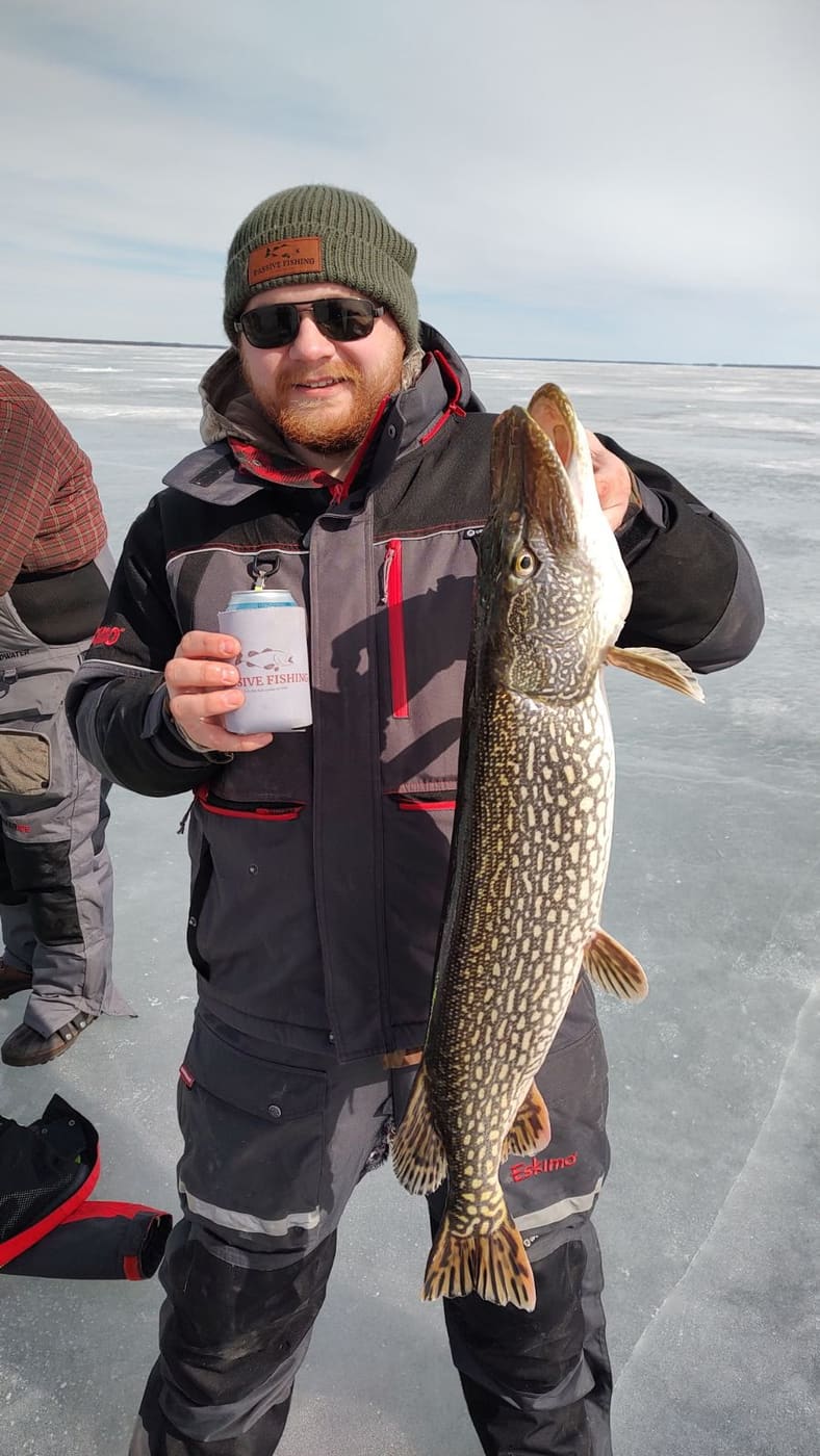 Angler in a Passive Fishing beanie holding a northern pike and a Passive Fishing koozie on the ice